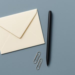 An overhead view of a plain cream envelope, a black pen and two paper clips on a steel blue surface.