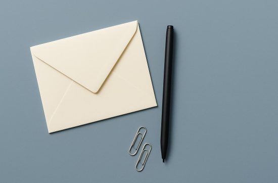 An overhead view of a plain cream envelope, a black pen and two paper clips on a steel blue surface.